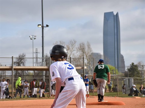 A base runner and a pitcher play on a field with Devon Tower in the background