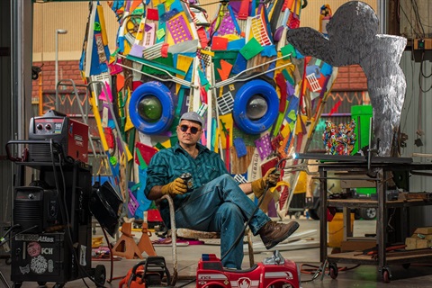 man holding blow torch sitting in chair with colorful sculpture behind him