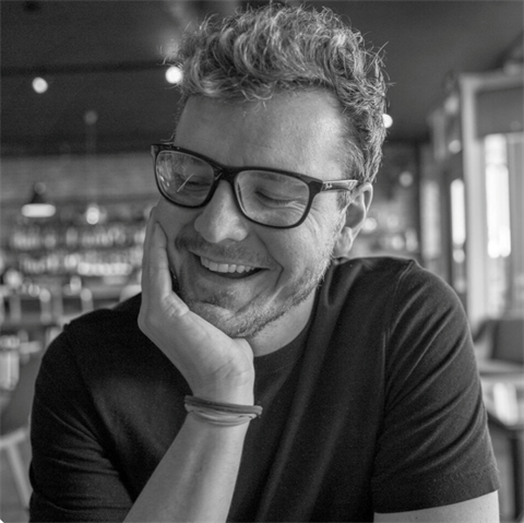 black and white photo of young white guy in black framed glasses smiling and resting head on hand