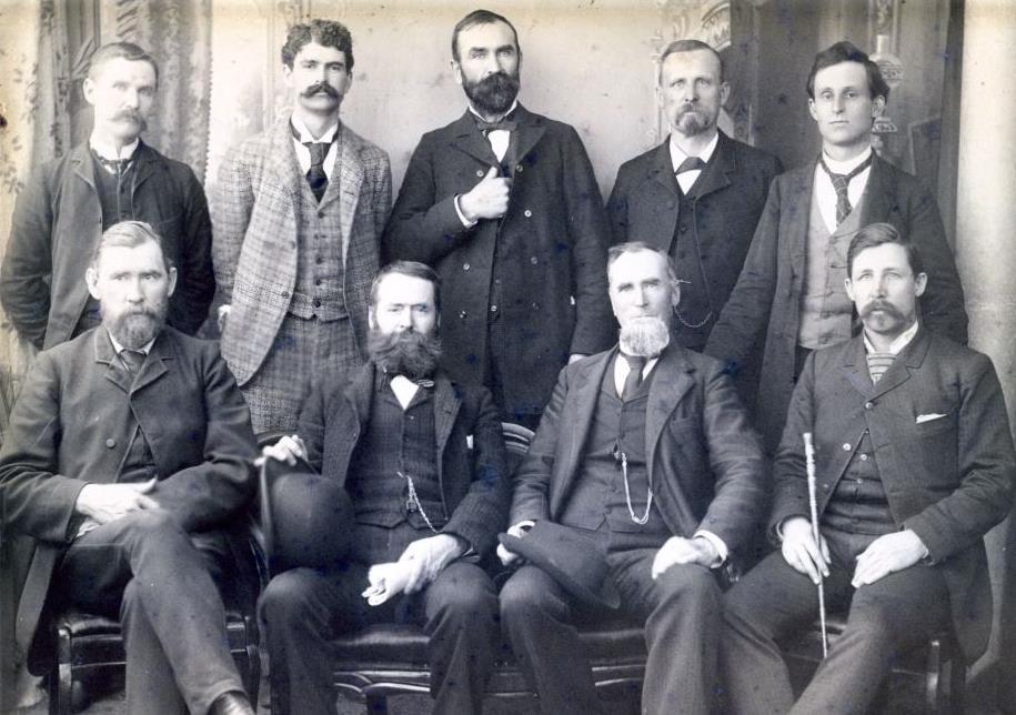 black and white photo of nine men wearing suits, four of whom are sitting down